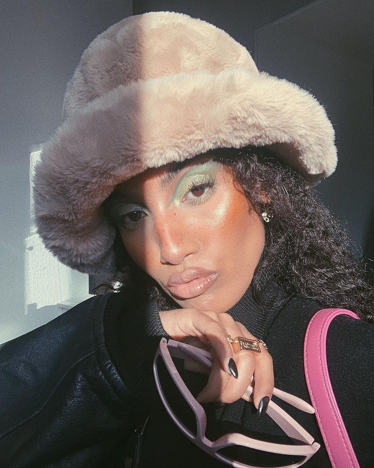 maquiagem-olho-verde-soblushing Garota usando maquiagem com sombra verde, chapéu de pelúcia e casaco preto. Ela está fazendo um bico com a boca e, com uma das mãos, segura um óculos de sol cor-de-rosa.