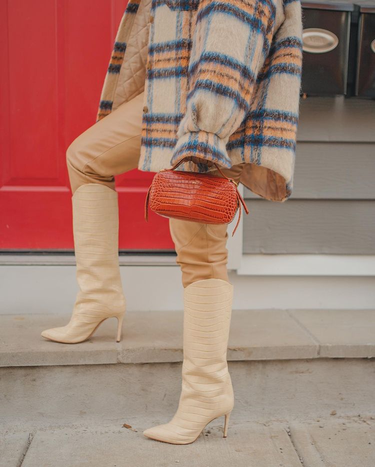 Na foto, aparece parte de uma camisa xadrez de flanela, calça bege e bota estilo cowboy com salto fino e textura croco também bege, além de uma bolsa laranja com textura croco segurada por uma das mãos da garota.