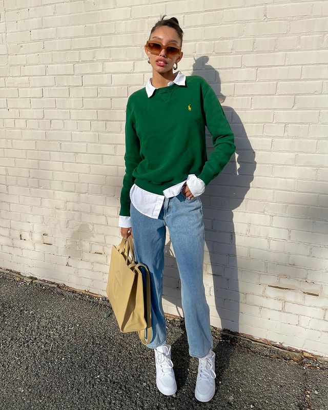 Garota usando camisa branca por baixo de suéter verde bandeira, calça jeans larguinha, tênis branco e uma das mãos segurando uma bolsa grande amarela-clara. Ela está em pé com uma das mãos no bolso.