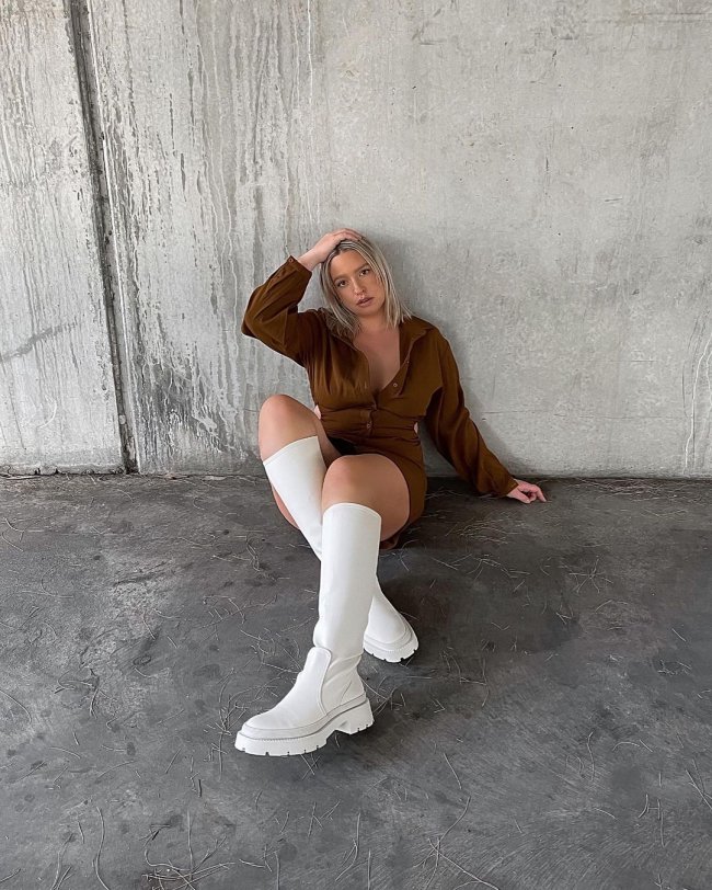 Mulher usando vestido marrom com mangas longas e bota até a altura do joelho off-white com sola tratorada. Ela está sentada no chão com uma mão na cabeça, a outra apoiada atrás e as pernas cruzadas.