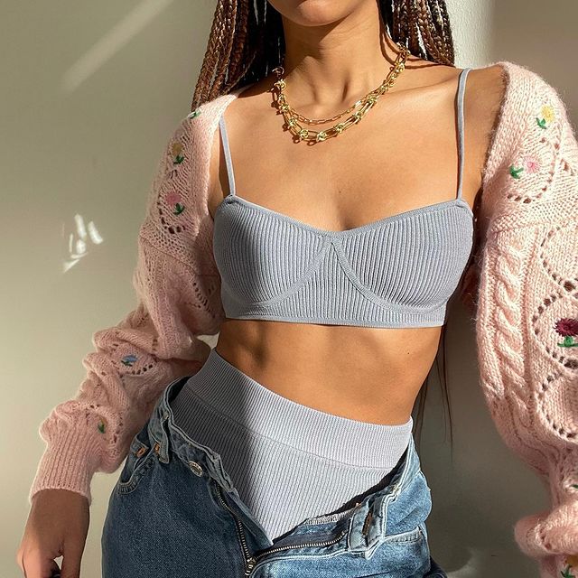 Garota usando top cropped azul pastel com hot pants da mesma cor. Ela está com uma calça jeans por cima desabotoada, além de um cardigan de tricô rosa com furinhos e flores bordadas e um colar de correntes douradas. Na foto, dá apenas para ver do pescoço para baixo até a altura do quadril.