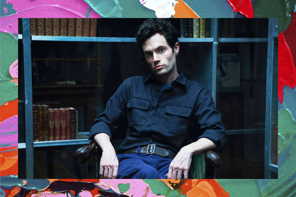 Penn Badgley, o Joe Goldberg em "You", sentado em uma cadeira em frente a uma estante de livros