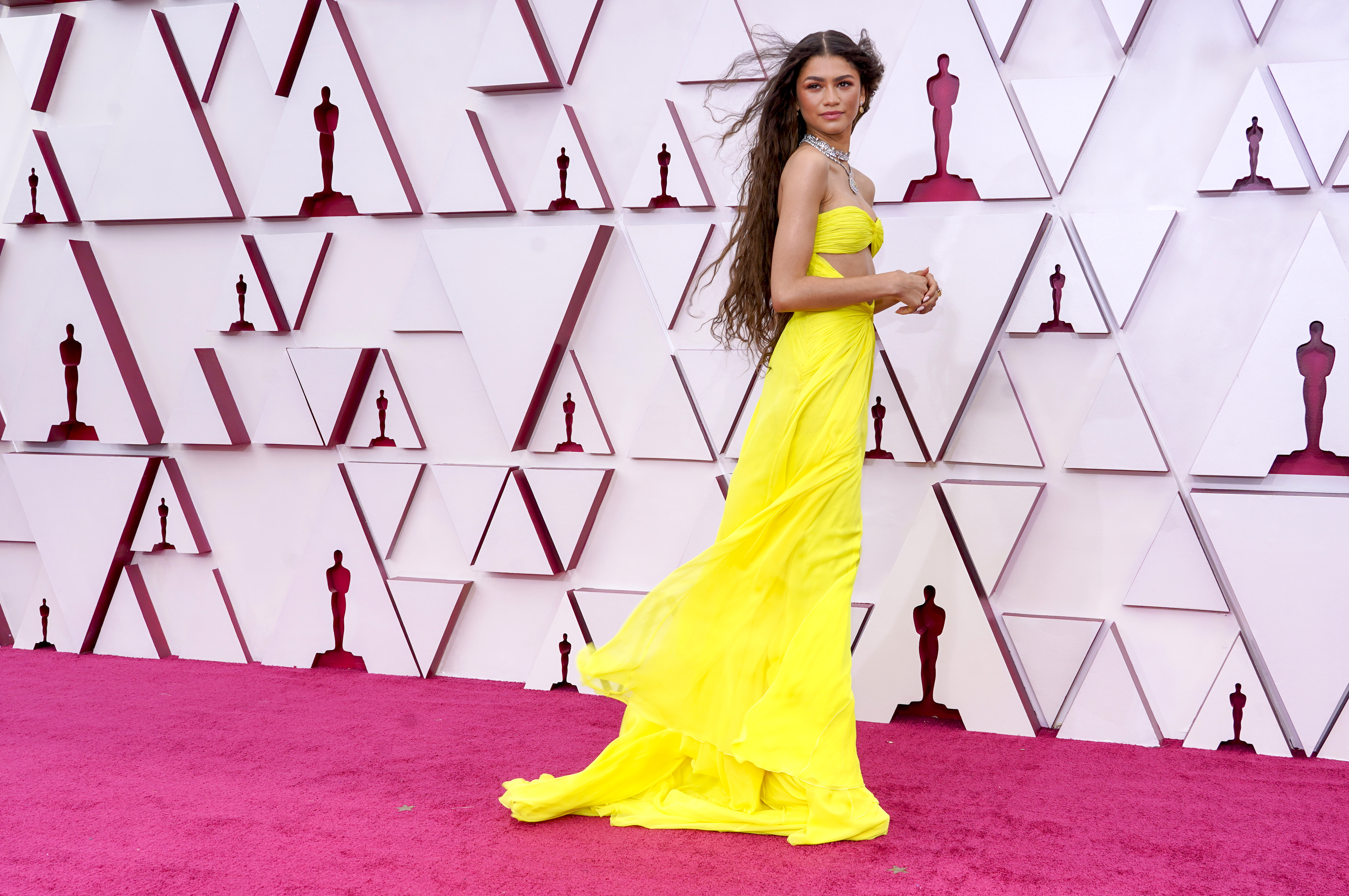 Zendaya usando vestido amarelo esvoaçante no Oscar 2021. Ela está com o corpo de lado, cabelo solto aos ventos, as mãos na frente do corpo e olhando para o lado com expressão facial séria.