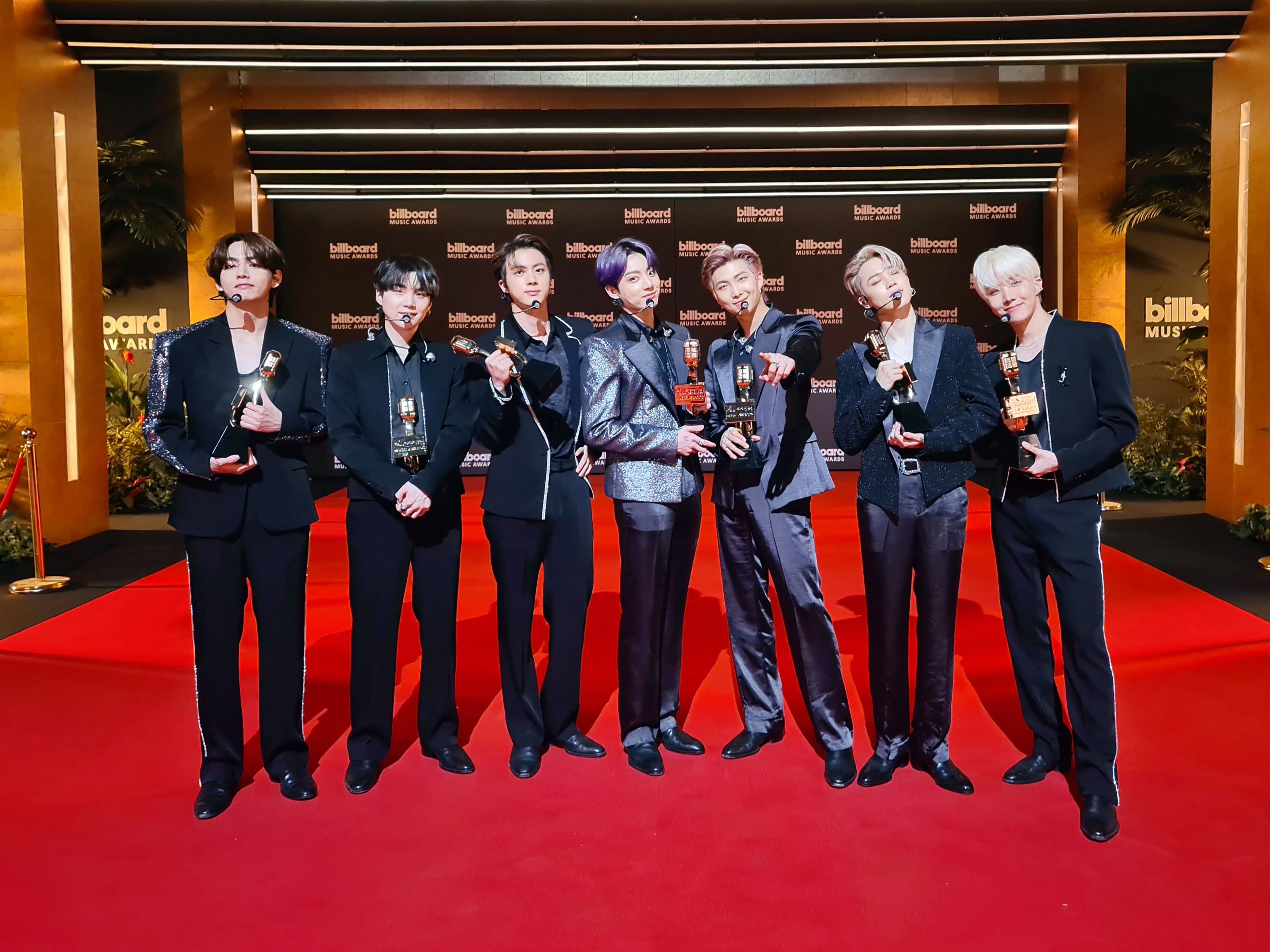 Sete integrantes do grupo BTS no red carpet do Billboard Music Awards 2021; eles estão alinhados na horizontal, cada um segurando um prêmio com roupas sociais escuras