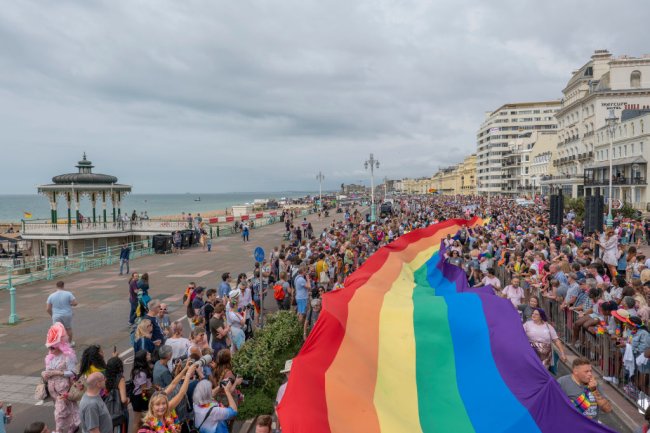 Foto aérea da Parada Gay que aconteceu em Brighton, cidade litorânea da Inglaterra, em 2019. Uma multidão carrega uma imensa banheira com as cores do arco-íris enquanto caminha.