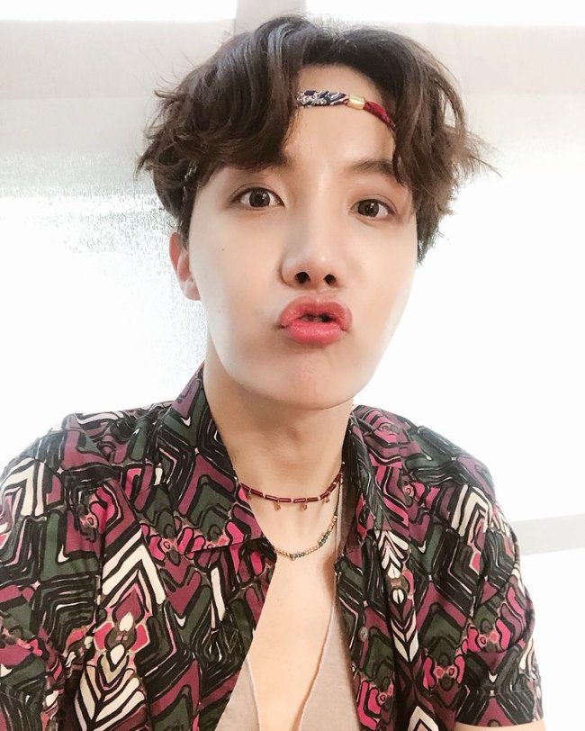 J-Hope BTS J-Hope, do grupo de k-pop BTS fazendo biquinho para foto, usando elástico na testa, colares e camisa estampada nos tons vermelho, verde, branco e preto.