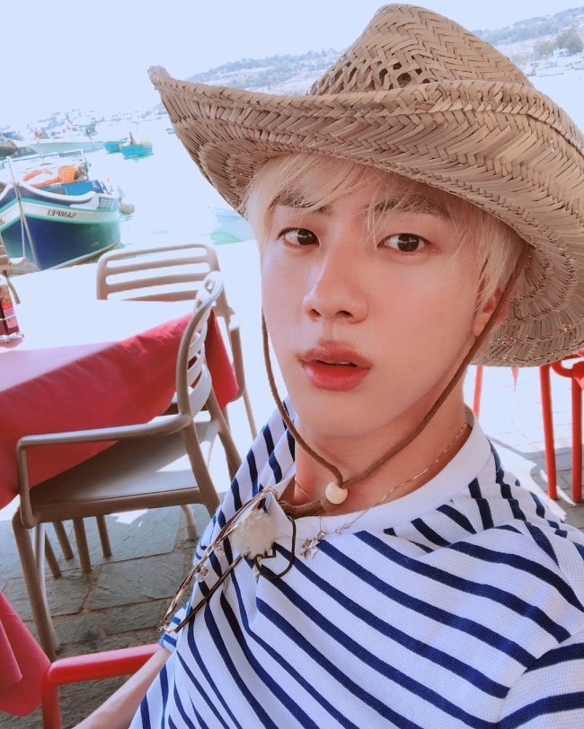 Jin, do grupo de K-pop, BTS sentado em uma cadeira branca de plástico, com expressão séria, usando chapéu de palha, camisa listrada e colares.