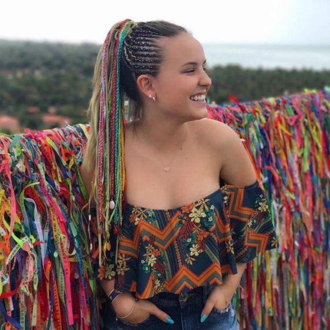 Larissa Manoela com cabelo longo olhando para o lado sorridente posando em frente a muro com fitinhas, usando cropped ciganinha com as mãos no bolso.