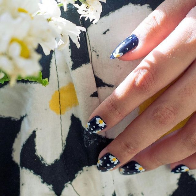 Nail art com flores e bolinhas Mão mostrando os dedos com esmalte azul e flor branca com detalhe amarelo ao lado de um tecido que parece o desenho da unha.