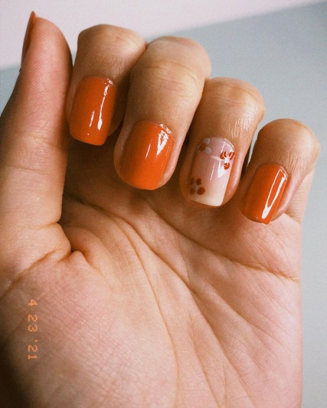 Nail art com florzinha laranja Mão com o dedo dobrado exibindo as unhas laranjas com uma com base clara e flores brancas e laranja.