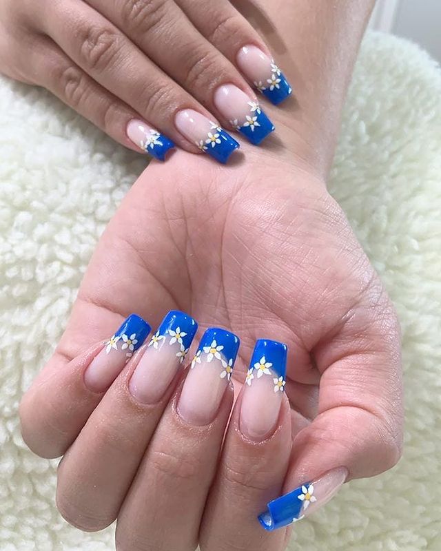 Nail art com francesinha azul e florzinha Duas mãos exibindo as unhas com base clara e francesinha azul com florzinha branca.
