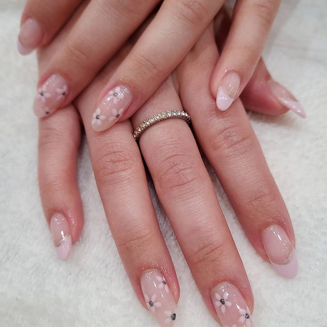 Nail art com francesinha delicada e florzinha Mãos uma sob a outra, com um anel fininho, exibindo as unhas com francesinha delicada em tom nude e pequenas florzinhas brancas.