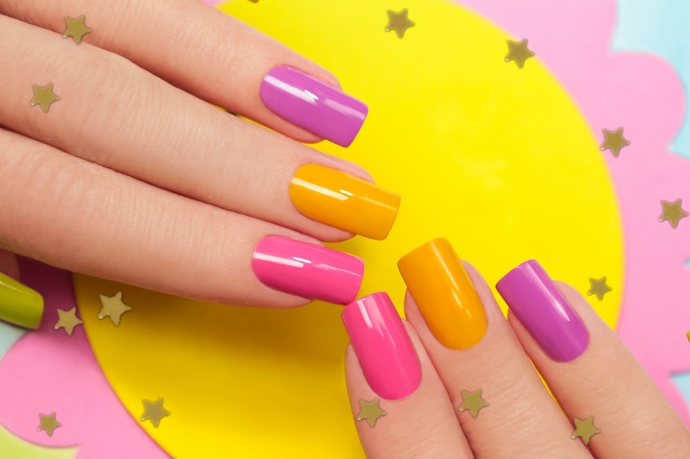 Unhas sendo exibidas em cima de um papel amarelo e outro rosa. As unhas são das cores: rosa, laranja e roxo.