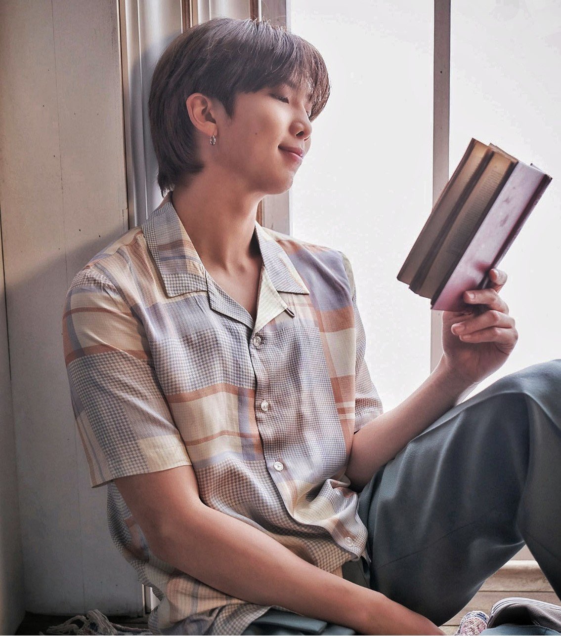 RM-livro2 RM do BTS sentado em uma janela lendo um livro, ele segura o livro com apenas uma das mãos e a outra está apoiada na perna