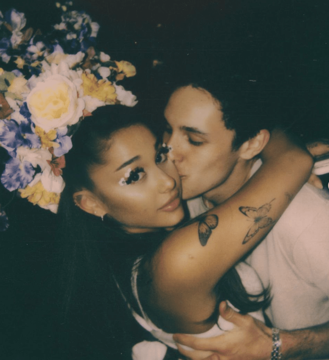 Foto de Ariana Grande e Dalton Gomez abraçados, ele está dando um beijo na bochecha dela, ela olha para câmera e está com os braços em volta do pescoço de Dalton em um abraço