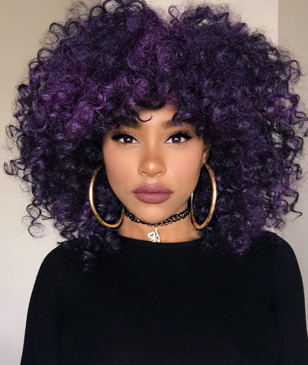 cabelo-colorido-roxo-cacheado Foto de uma mulher. Ela está vestindo uma blusa de manga longa preta, brinco de argola dourado, chocker preta e batom marrom. O cabelo dela é roxo escuro, ela olha para a câmera mas não sorri.
