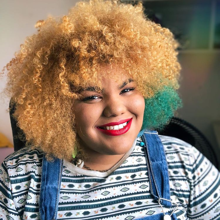 cabelo-crespo-mecha-lateral-azul Foto de uma mulher sorrindo para a câmera. Ela está vestindo uma blusa com estampa de listras e uma jardineira jeans, usa batom vermelho e tem o cabelo crespo ruivo com uma mecha na lateral esquerda na cor azul.