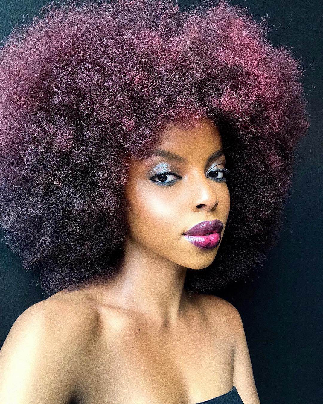 cabelo-crespo-pontas-roxas Selfie do rosto e cabelo de uma mulher. Ela está com um batom roxo e maquiagem metalizada nos olhos. O cabelo dela está solto, ele é crespo e tem as pontas na cor roxa
