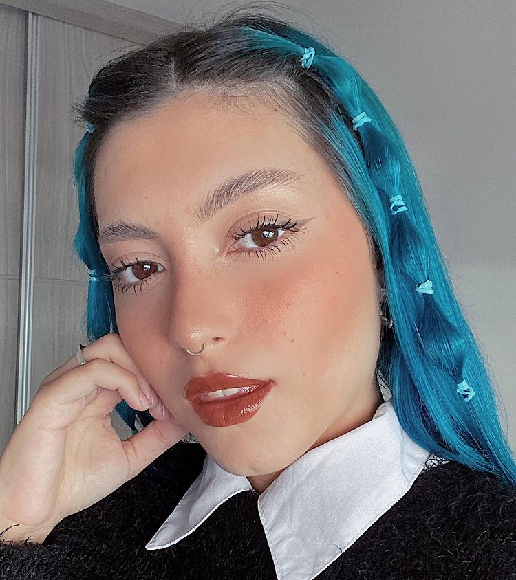 cabelo_azul_penteado_franja Selfie de uma mulher. Ela está usando uma camisa branca com um casaco de sobreposição, cabelo azul com a franja presa com elásticos coloridos. Ela olha para a câmera, não sorri e apoia o rosto na mão direita.