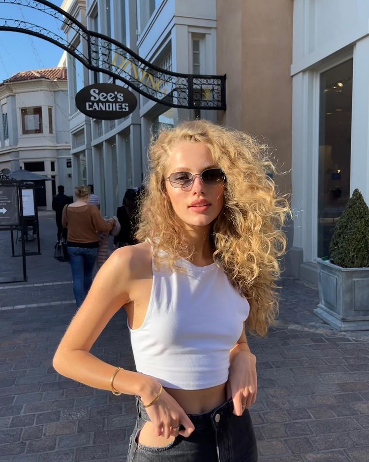 Foto de uma mulher na rua. Ela usa um top cropped branco, calça preta e está com o cabelo ondulado solto. Ela olha para a câmera e não sorri.