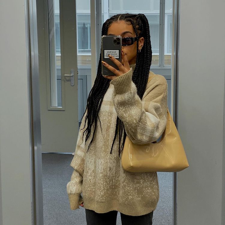 cabelo_maria_chiquinha_tranças Foto de uma mulher na frente do espelho. Ela está usando calça preta, casaco de frio bege com marrom, bolsa bege e cabelo com tranças preso em um penteado simples de maria chiquinha.