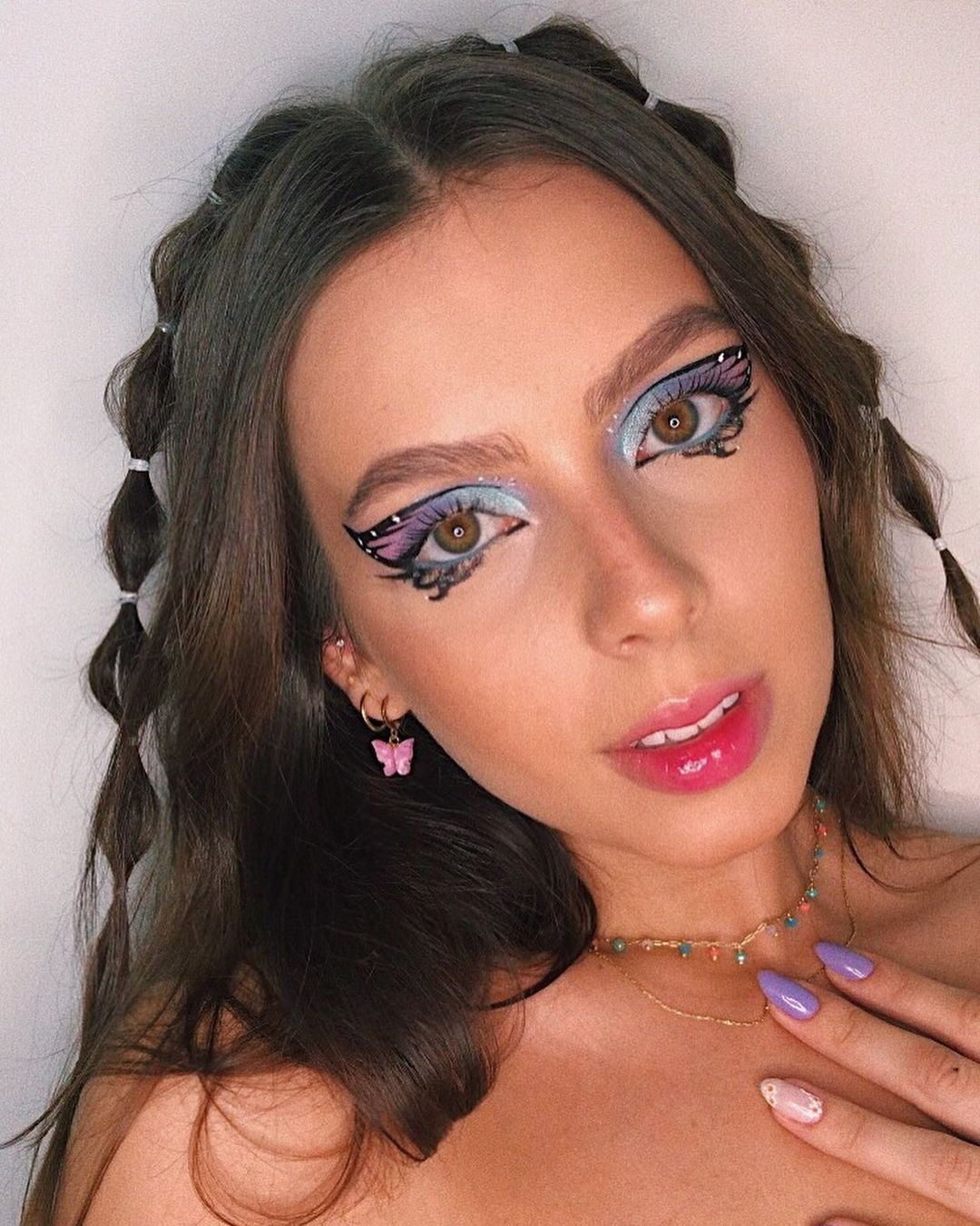 cabelo_penteado_bolha Selfie de uma mulher. Ela usa um colar dourado com pingentes coloridos, maquiagem artística de borboleta e penteado simples no estilo bolha. Ela olha para a foto mas não sorri.