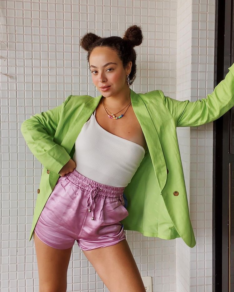 cabelo_penteado_coque_duplo Foto de uma mulher na varanda de uma casa. Ela veste um shorts rosa, body branco com manga única, casaco verde e penteado de coque duplo. Ela faz pose apoiando a mão direita na cintura e a esquerda na parede.