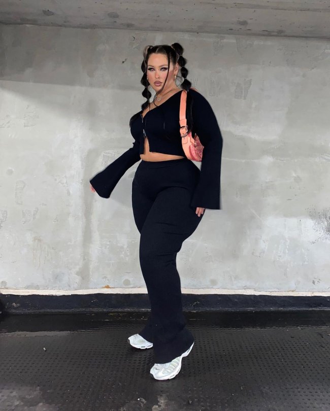 Foto de uma mulher na frente de uma parede branca. Ela usa um cardigan preto, calça legging bailarina preta, tênis branco e sholder bag rosa. Ela olha para a câmera e não sorri.