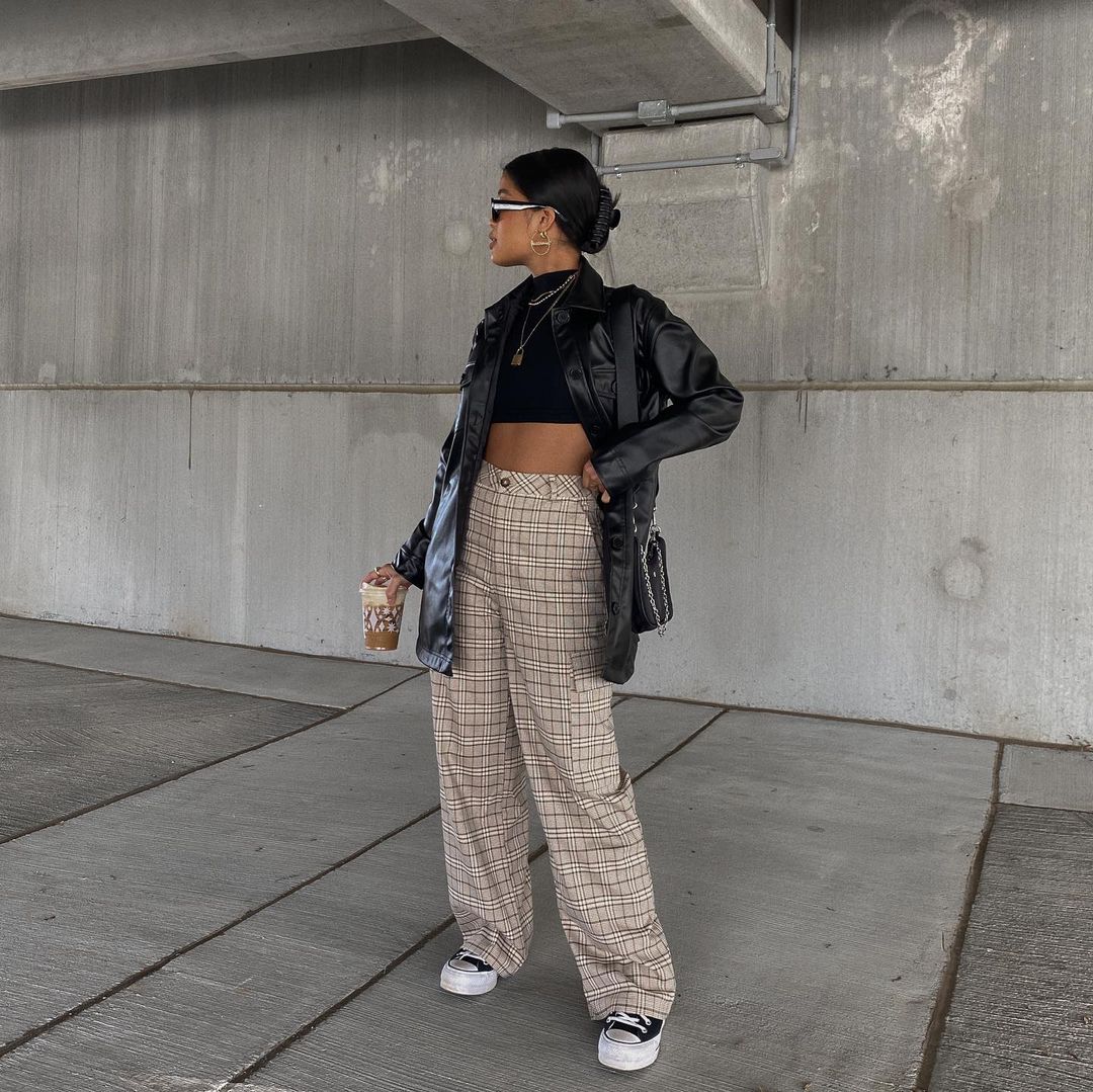 Foto de uma mulher em um estacionamento. Ela usa um cropped preto, jaqueta de couro preta, calça xadrez branca com marrom, tênis preto e bolsa preta. Ela olha para o lado e segura um copo de café.