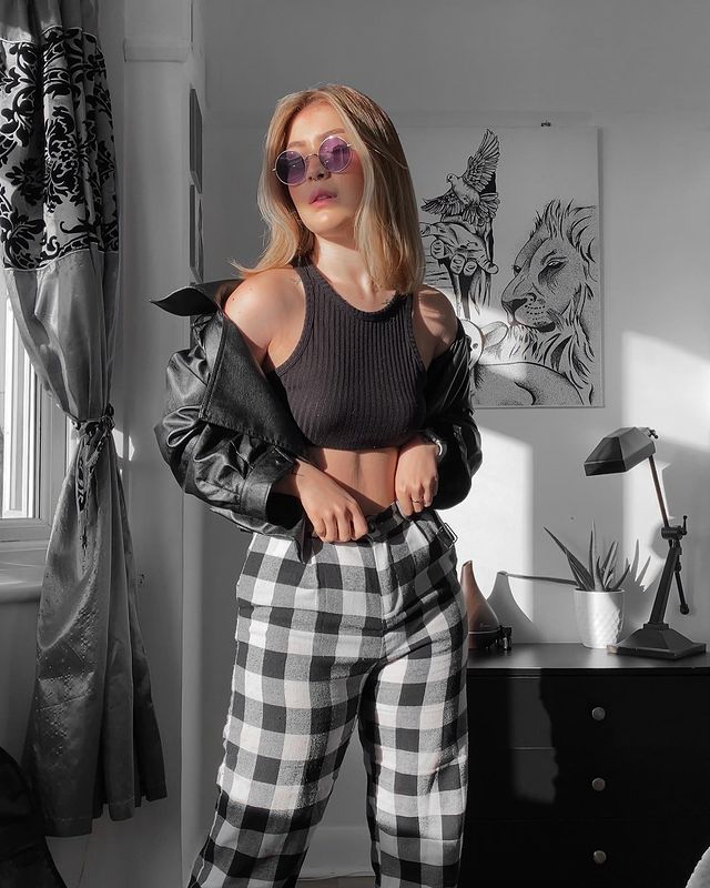 calca_xadrez_jaqueta_de_couro Foto de uma mulher em um quarto. Ela usa um cropped preto, calça xadrez preta e branca e jaqueta de couro preta. Ela olha para o lado e não sorri.