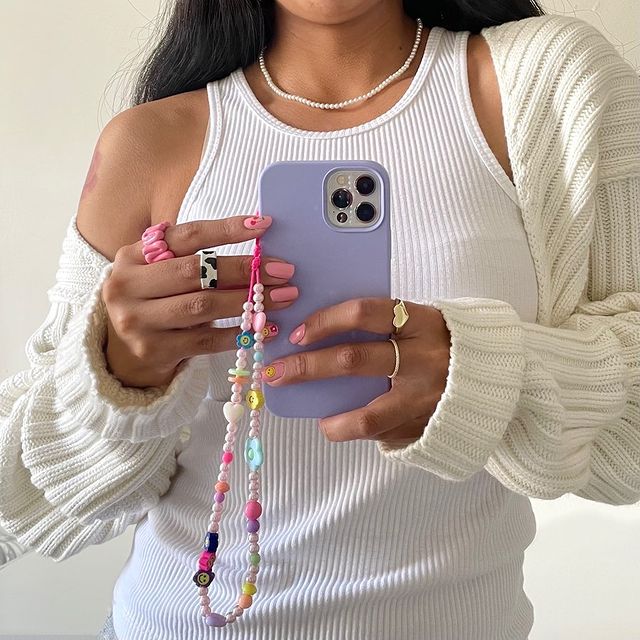 Garota usando regata branca, cardigan off white e tirando foto na frente do espelho segurando o celular que tem uma cordinha de miçangas pendurada. Na foto, só dá para ver da cintura até a altura do pescoço.