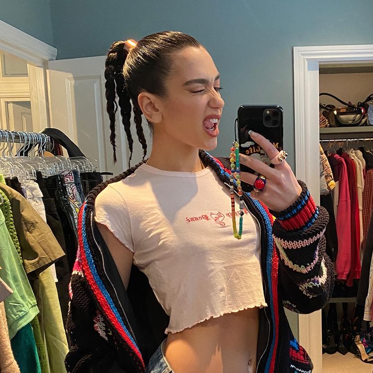 Dua Lipa usando top cropped branco e cardigan colorido em frente ao espelho. Ela está de lado, segurando o celular que tem uma cordinha de miçangas pendurada, e fazendo uma careta piscando o olho e mostrando a língua.