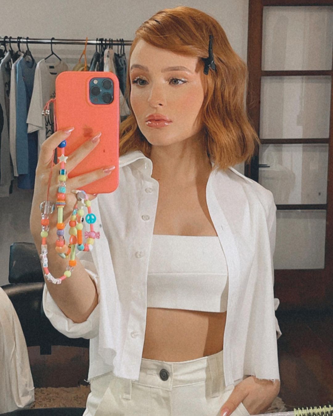 cordinha-celular-larissa-manoela Larissa Manoela usando conjuntinho todo branco formado por top cropped, calça e camisa. Ela está em frente ao espelho, segurando o celular que tem uma cordinha de miçangas pendurada, e com expressão facial séria.