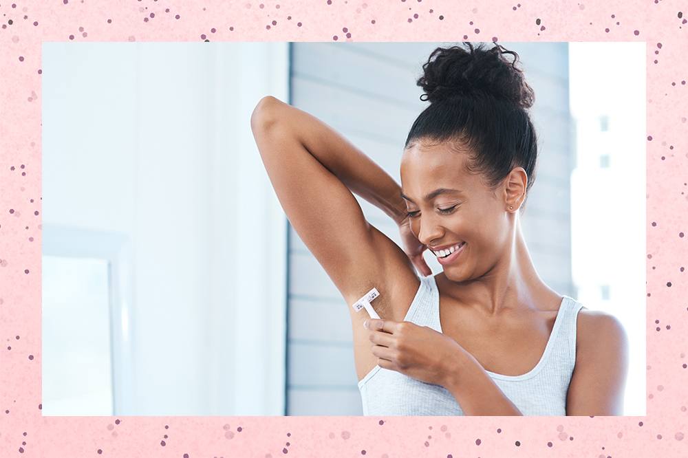 depilacao-axila-skincare No banheiro, uma garota está usando uma regata branca, com o cabelo preso em um coque, enquanto levanta um dos braços e faz depilação com lâmina na axila. Ela está olhando em direção à axila e sorrindo. O fundo da montagem é rosa com bolinhas.