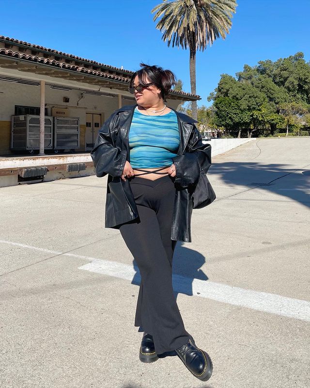 lauren_calca_legging_bailarina Foto de uma mulher na rua. Ela usa uma jaqueta de couro preta, blusa azul, calça legging bailarina, coturno e um óculos de sol vintage. Ela está com as mãos na cintura e olha para o lado.
