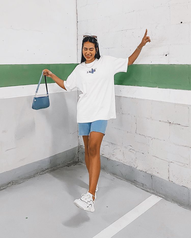 Foto de uma mulher em um estacionamento. Ela usa uma camiseta oversized branca, bermuda jeans, tênis branco e shoulder bag azul. Ela faz pose com a língua para fora e está de olho fechado.