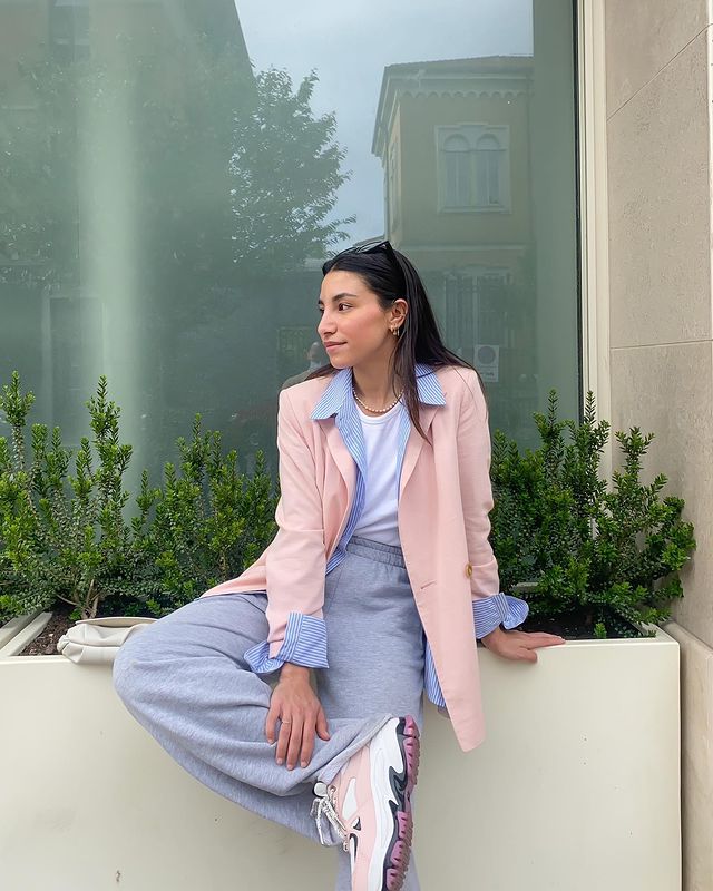 Foto de uma mulher sentada em um vaso de plantas grande. Ela usa uma camiseta branca, blazer rosa com azul, calça moletom cinza e tênis branco com rosa. Ela olha para o lado e sorri.