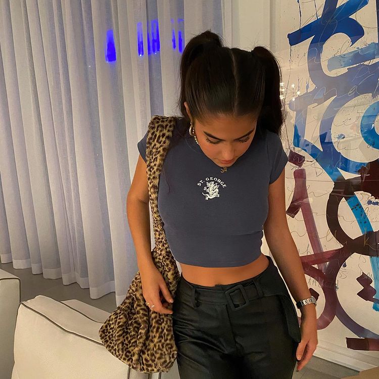 maria_chiquinha_penteado Foto de uma mulher em uma sala de estar. Ela veste um cropped azul, calça preta de couro, bolsa fluffy com estampa animal print e penteado maria chiquinha alto. Ela olha para o chão.