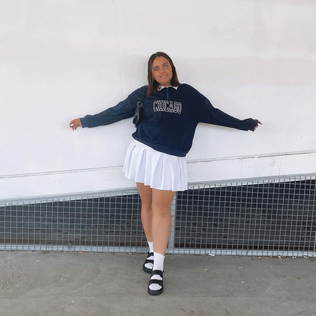 moletom_vintage_saia_colegial Foto de uma mulher com o fundo de uma parede branca. Ela usa um moletom vintage azul escuro, saia colegial branca e papete preta com meia branca. Ela olha para a câmera e sorri.