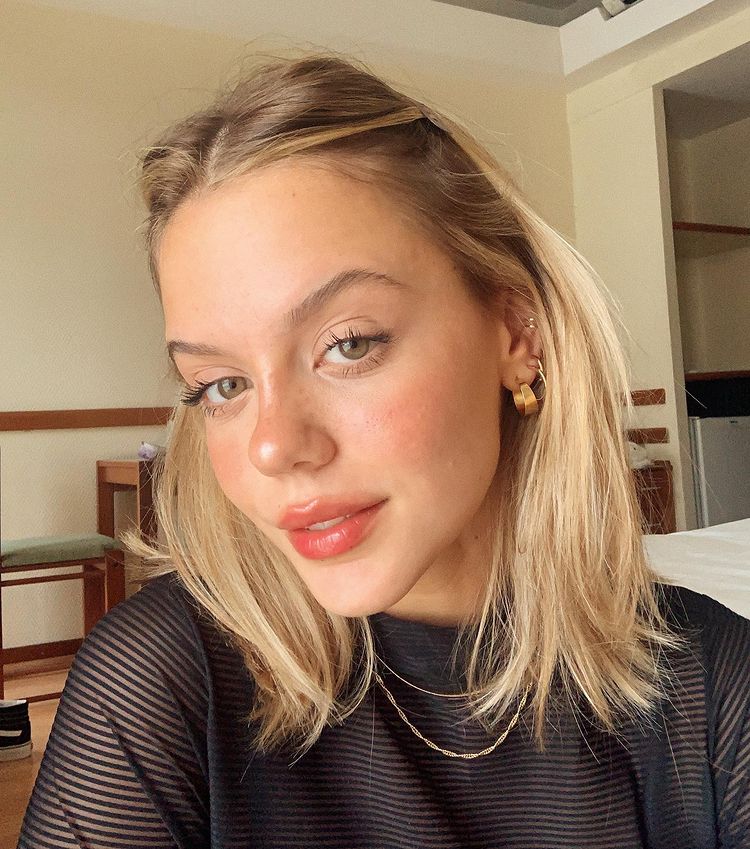 nic_cabelo_loiro Selfie de uma garota. Ela usa blusa de gola alta preta, brincos dourados e com a franja presa em um penteado. Ela olha para a câmera e não sorri.