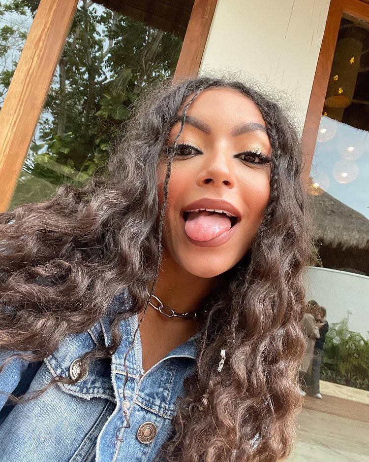 penteado_tranca_franja Selfie da Any Gabrielly. Ela está usando uma jaqueta jeans, colar prata, cabelo ondulado solto com um penteado de trança na franja com elástico. Ela olha para a câmera e mostra a língua.