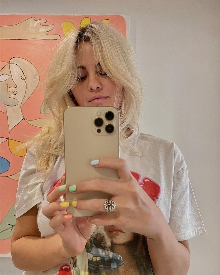 Selfie no espelho da cantora e atriz Selena Gomez. Ela usa uma camiseta branca com estampa vermelha, segura o celular com as duas mãos e está com o cabelo solto. Ela olha para o celular e não sorri para a foto.