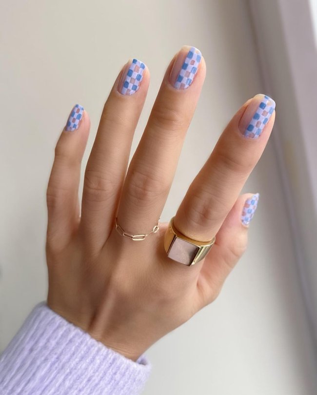 unhaxadrez Foto de uma mão com anéis no dedo médio e indicador. A unha está com uma nail art xadrez azul.