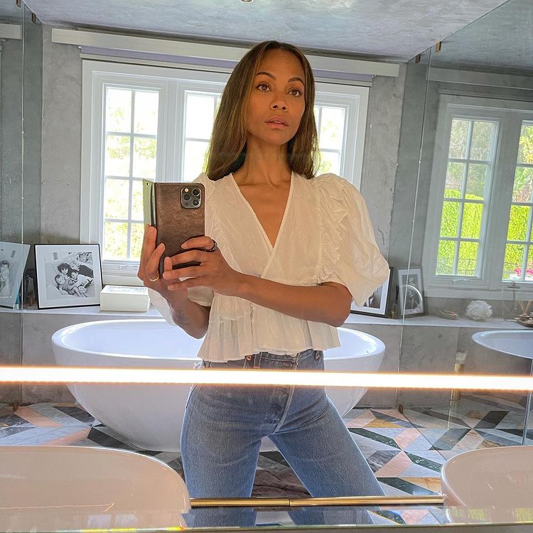 Selfie no espelho de Zoe Saldana; ela está no banheiro usando calça jeans clara e blusa branca; ao fundo é possível ver uma banheira e janelas refletindo a luz do dia; a expressão da atriz é mais séria e a foto está sendo tirada de um celular com capinha escura