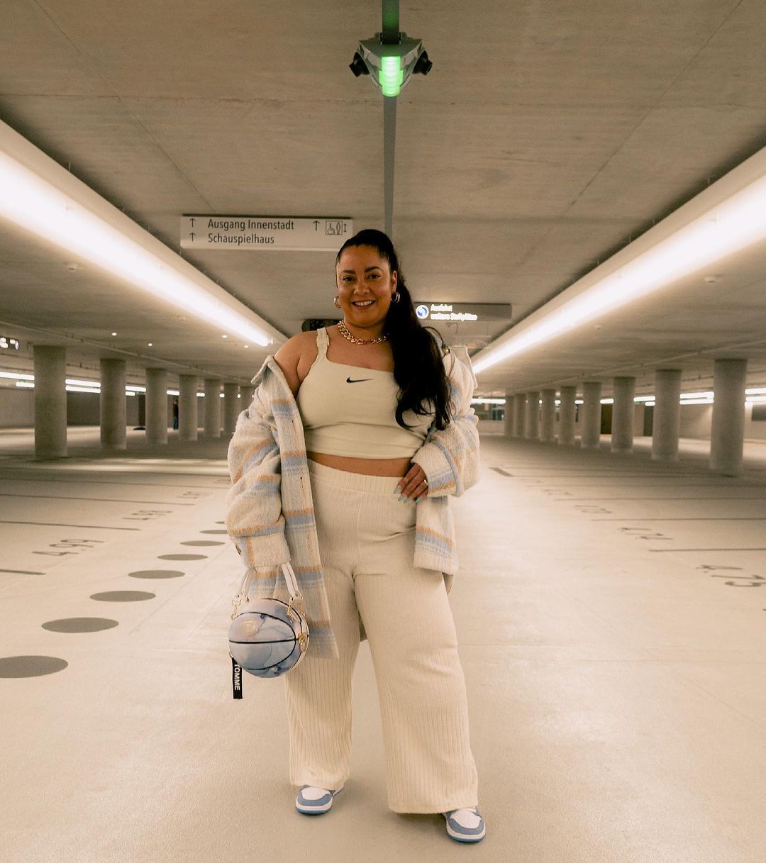 Foto de uma mulher no estacionamento. Ela usa um top e uma calça na cor creme, tênis nas cores azul e branca e segura a bolsa em formato de bola de basquete na mão direita. Ela olha para a câmera e sorri.