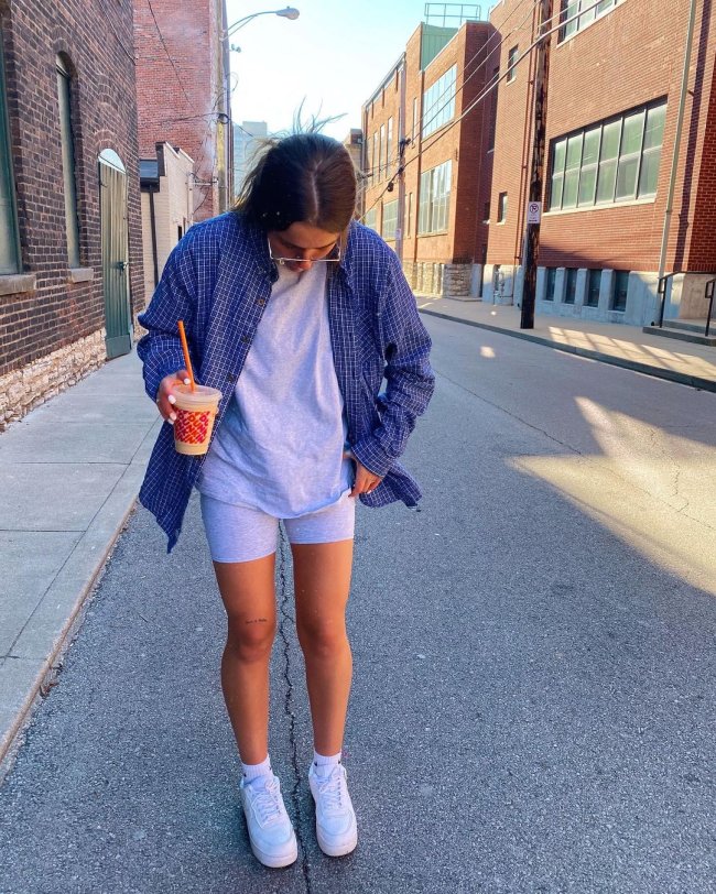 Foto de uma menina na rua. Ela usa uma camiseta cinza oversized, biker shorts, camisa xadrez azul e tênis branco. Ela olha para baixo e segura um copo de café.