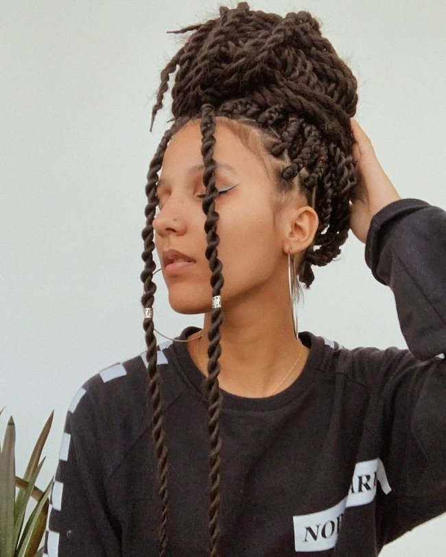 Jovem posando olhando para o lado com tranças por todo o cabelo formando um coque no topo da cabeça. Sua expressão é séria.