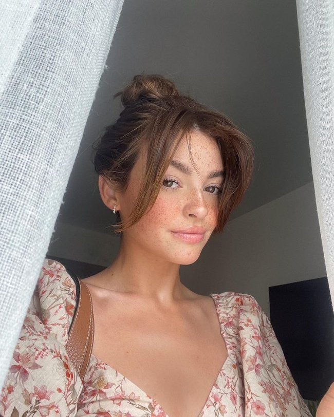 curtain bangs Selfie de uma mulher em uma janela. Ela usa um vestido floral e cabelo preso em um coque com franja cortininha solta. Ela olha para a câmera e sorri levemente.