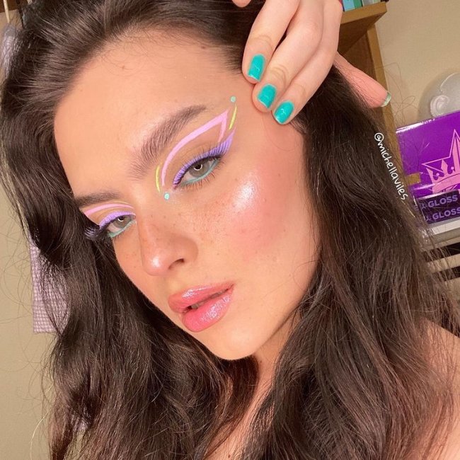 delineado gráfico tons pastel Selfie de uma mulher. Ela está com o cabelo solto, pele com aspecto natural e delineado gráfico em formato de borboleta em tons pastel. Ela segura o rosto com a mão direita, olha para a câmera e não sorri.