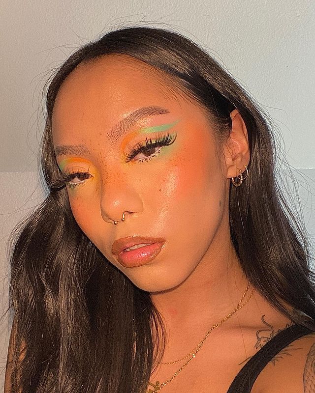 delineado pastel Selfie de uma mulher. Ela usa uma blusa de alcinha preta, cabelo solto, brincos de argola, maquiagem com pele natural e delineado gráfico em tons de amarelo, laranja e verde pastel. Ela olha para a câmera e não sorri